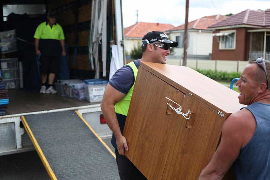 Two professional movers team up to lift a heavy cabinet down a moving truck ramp.